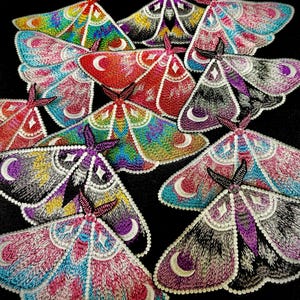May include: A collection of embroidered moth patches in various colors, including pink, blue, green, and purple. Each patch features intricate details, such as crescent moon and star designs, and is set against a black background.
