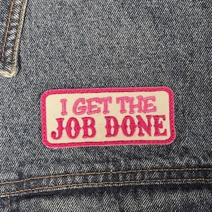 Könnte beinhalten: Ein rechteckiger, bestickter Aufnäher auf blauem Jeansstoff. Der Aufnäher hat einen cremefarbenen Hintergrund mit einem leuchtend rosa Rand und dem Text I GET THE JOB DONE, der in fetten, rosa, dekorativen Buchstaben in der Mitte gestickt ist.