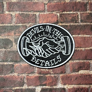May include: Black and white embroidered patch with the text "Devils in the Details" in a circle. The patch features a handshake design.