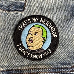 Puede incluir: Un parche redondo bordado con un personaje de dibujos animados con pelo verde y la boca abierta, sobre un fondo azul claro. El parche está bordeado por un anillo negro con el texto "THAT'S MY NEIGHBOR I DON'T KNOW YOU!"