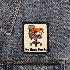 Könnte beinhalten: Ein rechteckiger gestickter Aufnäher auf einer blauen Jeansjacke. Der Aufnäher hat einen cremefarbenen Hintergrund mit schwarzem Rand und zeigt eine orangefarbene Garnele in einem grauen Bürostuhl mit dem Text My Back Hurts in Schwarz.