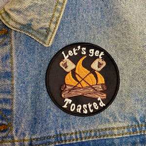 May include: A black embroidered patch with a campfire, marshmallows, and the text "Let's get toasted".