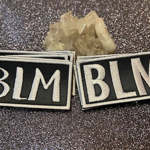 May include: A stack of black fabric patches with white embroidered letters spelling out "BLM".