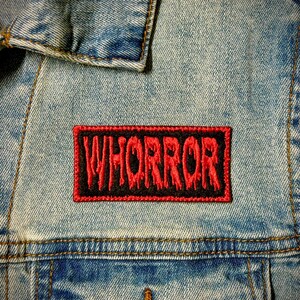 Peut inclure: Un écusson rouge et noir avec le mot "WHORROR" en lettres rouges qui coulent cousu sur une veste en jean bleue.