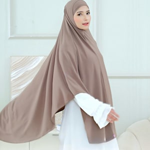 Può includere: Un hijab color tortora caratterizzato da un tessuto liscio e drappeggiato che copre la testa e le spalle, indossato sopra un indumento bianco a maniche lunghe con polsini a volant. Il velo si estende sul retro e sul lato, creando una silhouette fluida.