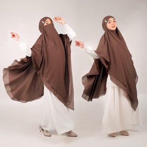 May include: Two women wearing brown hijab and white outfits pose against a white backdrop. The hijab drapes over the shoulders and covers the head and face. The outfits include long sleeves and wide-legged pants. The fabric appears lightweight and flowing.