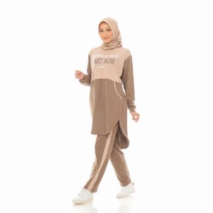 May include: A two-piece outfit consisting of a long-sleeved tunic and matching pants in shades of beige and brown. The tunic features a high neckline, a hood, and the text "START NOW" printed on the chest. The pants have a side stripe.