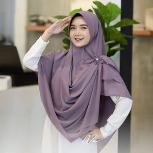 May include: A lilac hijab with a flower pin, worn over a white long-sleeved shirt. The hijab is draped with soft folds and a decorative flower pin. The image shows a woman smiling and saluting.
