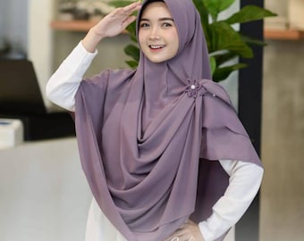 May include: A lavender hijab with a flower pin, draped over a white long-sleeved shirt. The hijab is styled with soft folds and a decorative flower pin. The image shows a woman smiling and saluting.