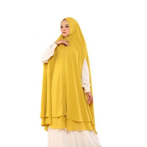 Può includere: Un hijab giallo senape con un design a strati. L'hijab è indossato sopra una camicia bianca a maniche lunghe e una gonna bianca. La modella è in piedi davanti a uno sfondo bianco.