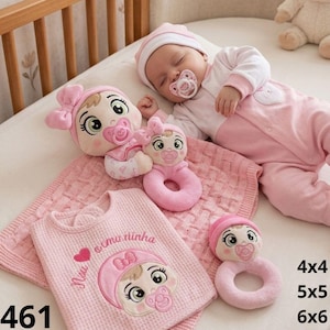 May include: A baby set featuring a pink waffle-knit blanket with an embroidered baby face and the text 'Nini é a minha', two pink plush baby rattles with embroidered faces, and a plush doll with a pink bow, all in shades of pink and white.