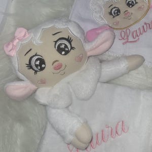 May include: A white plush lamb toy with pink ears, a pink bow, and embroidered eyes. The lamb is holding a white blanket with the name "Laura" embroidered in pink.