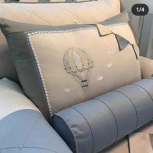 May include: A decorative pillow with an embroidered hot air balloon design. The pillow features a light beige background with a blue border and white trim. Additional pillows in blue and white are also visible.