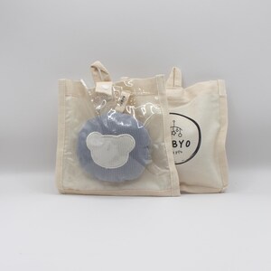 May include: A light beige tote bag with a clear front pocket holding a blue plush toy with a white bear face. A second beige tote bag is visible with a black and white logo and the letters "BYO".