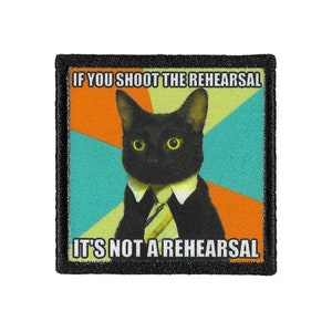 Puede incluir: Un gato negro con traje y corbata con el texto "Si grabas el ensayo, no es un ensayo" en un parche.