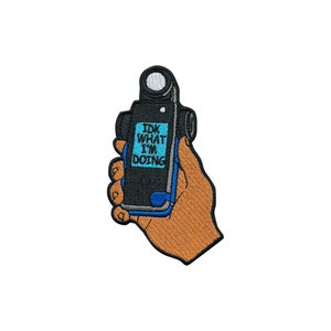 Puede incluir: Parche bordado para planchar con una mano sosteniendo una cámara con el texto "IDK WHAT I'M DOING" en la pantalla. La cámara es negra con una pantalla azul y una lente plateada. La mano es marrón.
