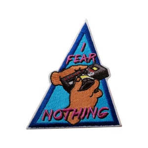 Puede incluir: Parche bordado en forma triangular con borde azul. El parche presenta un oso de dibujos animados sosteniendo un arma, con las palabras "I FEAR NOTHING" en letras rosas. El oso es marrón y el arma es negra.