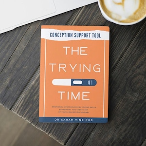Op de afbeelding: Een oranje boek getiteld "The Trying Time" met een blauw en wit zwangerschapstestpictogram. De ondertitel van het boek is "Emotionele en psychologische copingvaardigheden die je ondersteunen bij elke stap van je conceptiereis". De auteur is Dr. Sarah Vine PhD.
