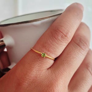 Può includere: Un delicato anello d'oro con una piccola gemma rotonda verde chiaro. L'anello ha una fascia sottile e un design semplice ed elegante. La gemma è incastonata in un'incastonatura a quattro griffe.