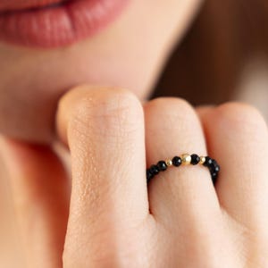 Könnte beinhalten: Ein zarter Ring aus schwarzen Perlen und goldenen Akzenten. Der Ring wird an einem Finger getragen.