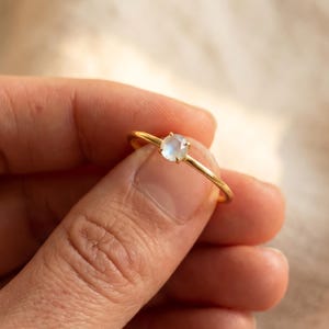 Op de afbeelding: Een gouden ring met een kleine, ronde, lichtblauwe edelsteen. De ring wordt in een hand gehouden tegen een zachte, neutrale achtergrond. De band is dun en delicaat, waardoor de edelsteen wordt benadrukt.