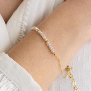 Peut inclure: Un bracelet doré délicat orné de petites perles blanches. Le bracelet est doté d'une fine chaîne et d'un fermoir dorés. Les perles sont espacées régulièrement le long du bracelet, avec de petits accents dorés. Le bracelet est porté sur un poignet.