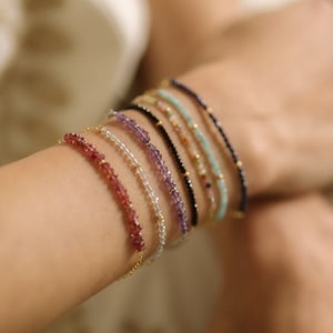 Può includere: Il polso di una donna adornato con una pila di braccialetti di perline colorate. I braccialetti sono fatti di varie pietre preziose, tra cui ametista, turchese e rubino.