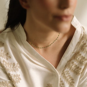 A fine gold necklace adorned with tiny, vivid green beads. The necklace is set against a cream blouse featuring gold floral embroidery. The image is well-lit, accentuating the jewellery's details and the blouse's texture.