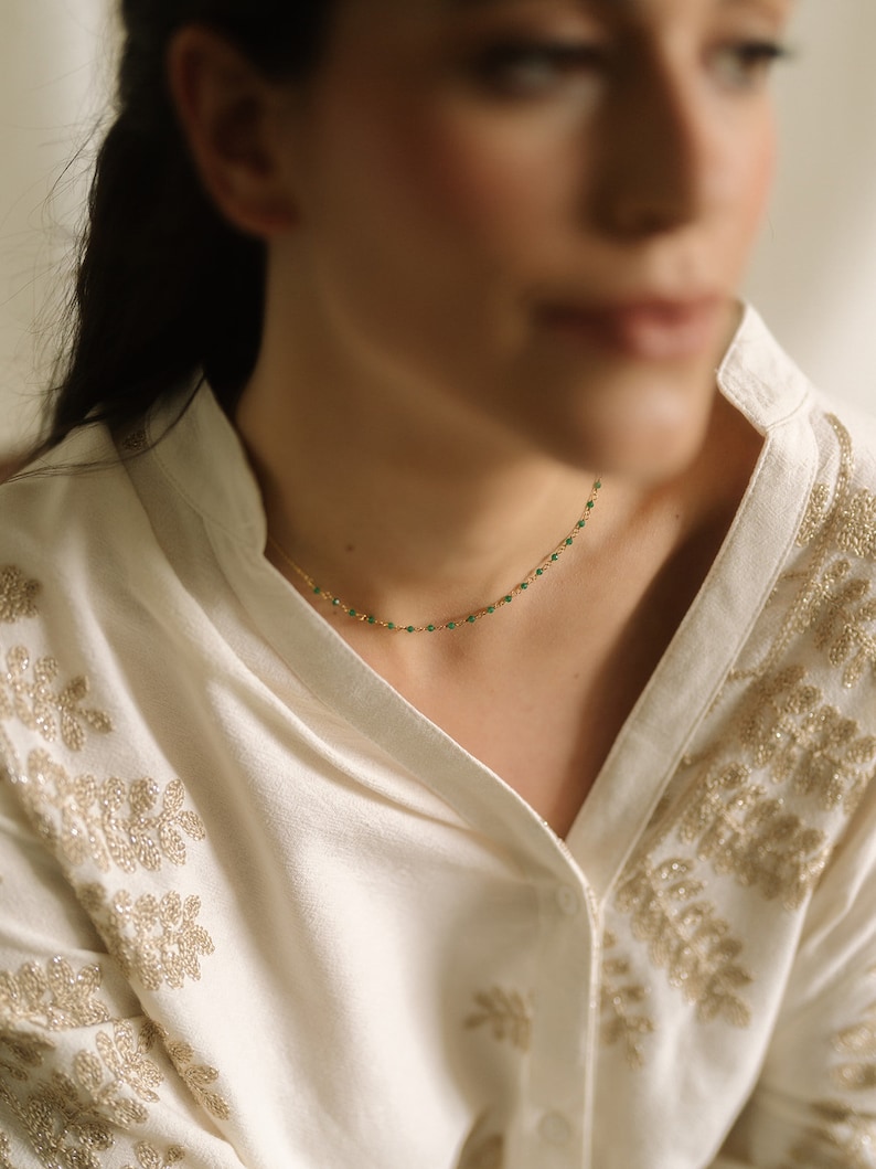A fine gold necklace adorned with tiny, vivid green beads. The necklace is set against a cream blouse featuring gold floral embroidery. The image is well-lit, accentuating the jewellery's details and the blouse's texture.