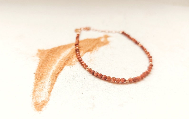 Dainty gemstone bracelet on a blanket of desert sand. The bracelet is made of tiny golden aventurine beads, in an orange color, and with rose gold accents on a white background. This women bracelet looks delicate and minimalist.