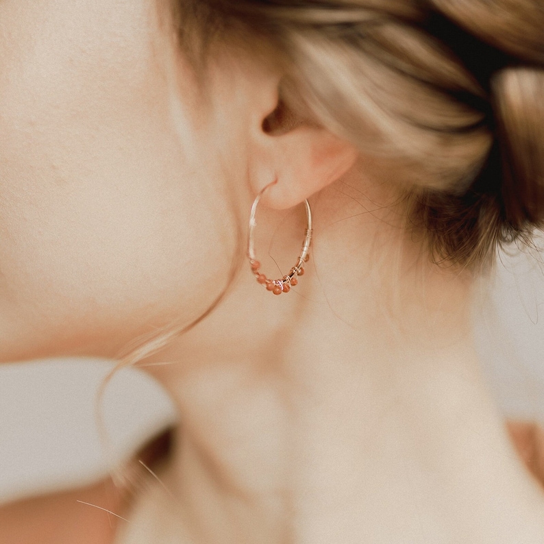 These golden aventurine hoop earrings are seen placed on the ear of a model, who is in profile. Their bohemian, minimalist feel is highlighted by the rose gold plating and tiny aventurine beads.