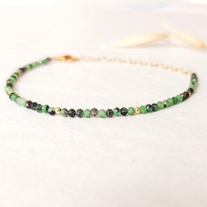 Peut inclure: Bracelet délicat orné de petites perles vertes et noires facettées, parsemées de perles dorées. Le bracelet est monté sur une fine chaîne dorée, créant un design minimaliste et élégant. Le bracelet est présenté sur une surface blanche.