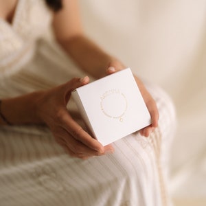 A white square box with gold lettering and a gold necklace illustration is held in a person's hands. The box has the word "ARENNA" printed on it. The person is wearing a white and beige striped dress.