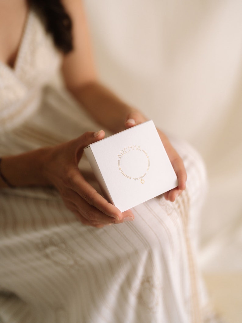 A white square box with gold lettering and a gold necklace illustration is held in a person's hands. The box has the word "ARENNA" printed on it. The person is wearing a white and beige striped dress.