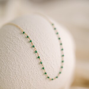 A fine gold necklace with small, vivid green beads strung along its length. The necklace is shown against a soft, textured white backdrop, accentuating the contrast between the gold and green.
