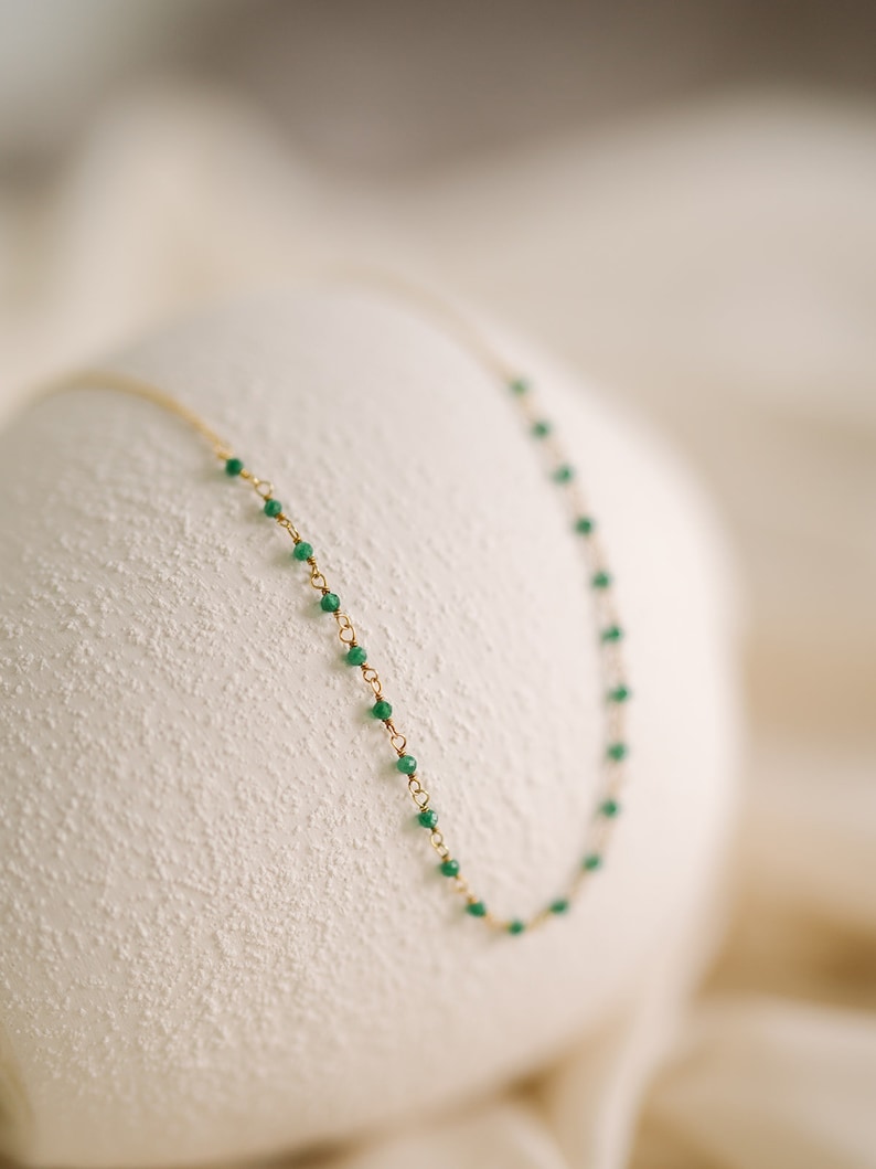 A fine gold necklace with small, vivid green beads strung along its length. The necklace is shown against a soft, textured white backdrop, accentuating the contrast between the gold and green.