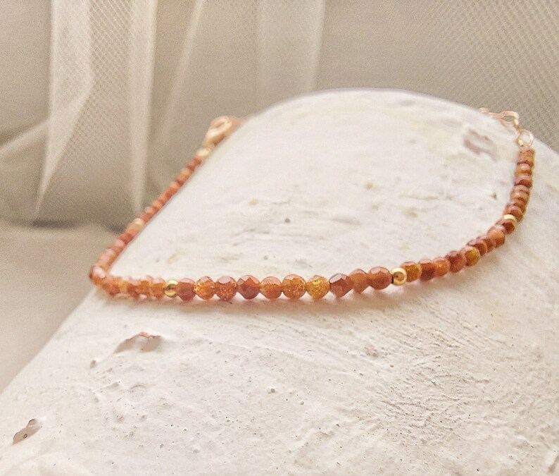 Tiny golden aventurine gemstones are seen up close on white surface. Rose gold accents and delicate rose gold chain look perfectly integrated with the aventurine beads. A few rose gold pieces can be seen among the aventurine stones.