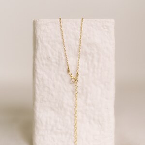 May include: A delicate gold-colored necklace with a chain and an adjustable length. The necklace is displayed on a white, textured block. The chain has a small clasp and a series of small loops for adjusting the length.