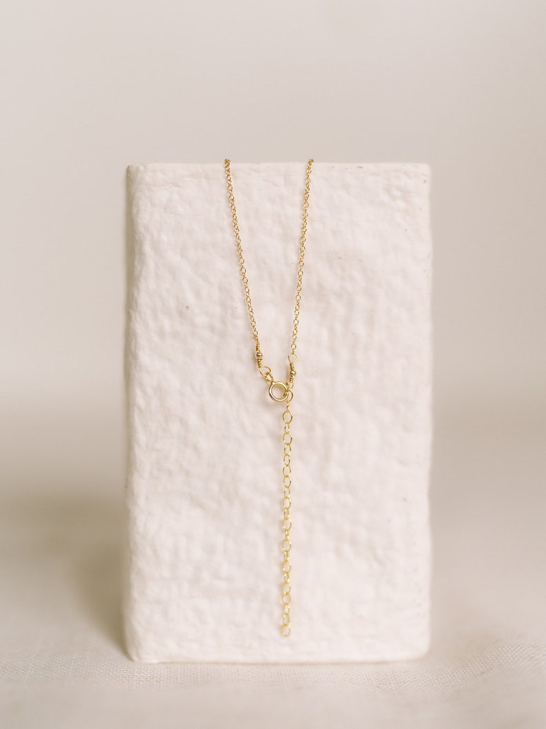 May include: A delicate gold-colored necklace with a chain and an adjustable length. The necklace is displayed on a white, textured block. The chain has a small clasp and a series of small loops for adjusting the length.
