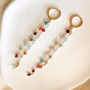 May include: Gold hoop earrings with beaded dangles in various colours. The beads are red, orange, yellow, green, blue, and purple. The earrings are presented on a white, curved surface.