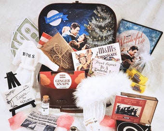 May include: A brown suitcase gift set themed around It's a Wonderful Life, containing a DVD, M&M's, ginger snaps, a miniature white bathrobe with BFHS, a small easel with a sign reading Lasso the Moon, a record player box, and movie-related props.