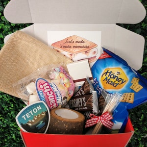 May include: A red box filled with s'mores making supplies. The box contains graham crackers, chocolate bars, marshmallows, skewers, a burlap bag, a brown paper card with the text "Let's make s'mores memories", and a sticker with the text "Grand Teton National Park".