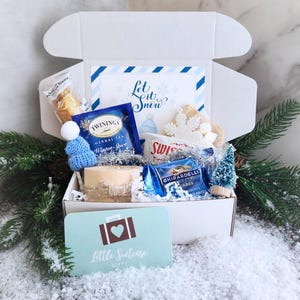 May include: A white gift box filled with winter-themed items, including a blue knit hat, tea, chocolate, and a card that says "Little Suitcase Gifts." The box also contains a card that says "Let it Snow."