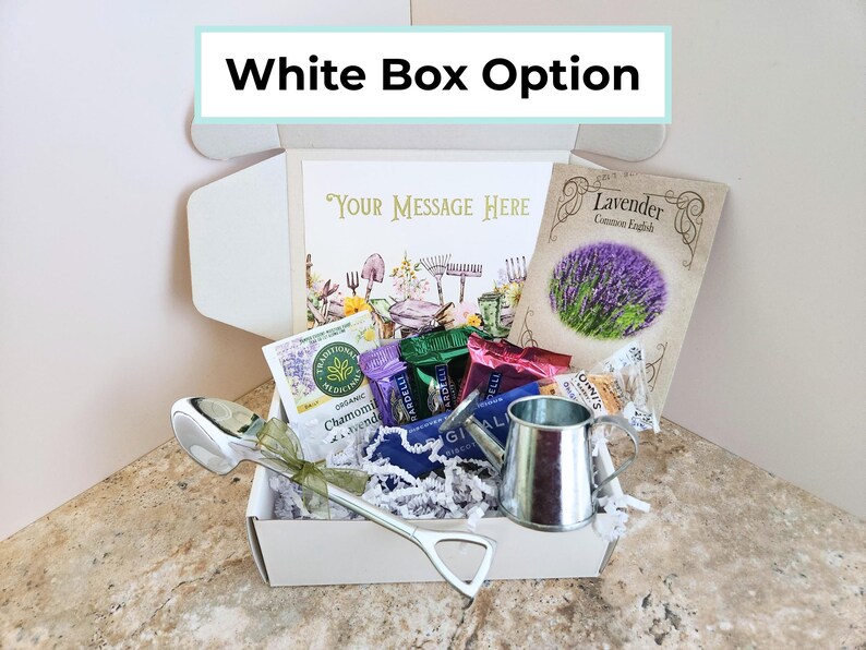 May include: A white gift box labeled "White Box Option" filled with various items. Inside are tea packets, a small watering can, a decorative spoon, and a packet of lavender seeds. A card with the text "Your Message Here" is also included.