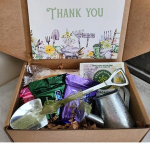 May include: A brown cardboard box filled with gardening-themed items. Includes a card with "Thank You" text, a miniature watering can, a shovel-shaped bottle opener, and various wrapped treats. The card features garden tools and floral illustrations.