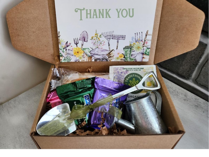 May include: A brown cardboard box filled with gardening-themed items. Includes a card with "Thank You" text, a miniature watering can, a shovel-shaped bottle opener, and various wrapped treats. The card features garden tools and floral illustrations.