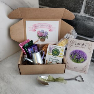 May include: A gift box with a "Happy Mother's Day" card, tea, lavender seeds, and a miniature watering can. The box contains wrapped candies and a decorative spoon with a green ribbon. The card features a watercolor of hydrangeas.