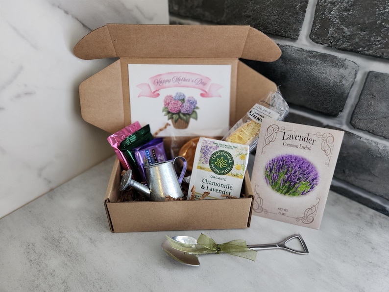 May include: A gift box with a "Happy Mother's Day" card, tea, lavender seeds, and a miniature watering can. The box contains wrapped candies and a decorative spoon with a green ribbon. The card features a watercolor of hydrangeas.