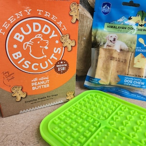 May include: A box of Buddy Biscuit dog treats with a gingerbread man design. The box says "Teeny Treat" and "Baked in the USA!"  The treats are made with natural peanut butter. A green silicone dog treat mat with a grid pattern. A bag of Himalayan Dog Chew with chicken flavor. The bag says "Naturally Healthy Dog Chew Chew Density - Hi".