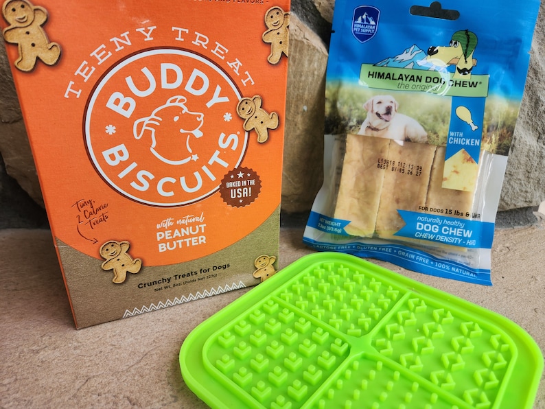 May include: A box of Buddy Biscuit dog treats with a gingerbread man design. The box says "Teeny Treat" and "Baked in the USA!"  The treats are made with natural peanut butter. A green silicone dog treat mat with a grid pattern. A bag of Himalayan Dog Chew with chicken flavor. The bag says "Naturally Healthy Dog Chew Chew Density - Hi".